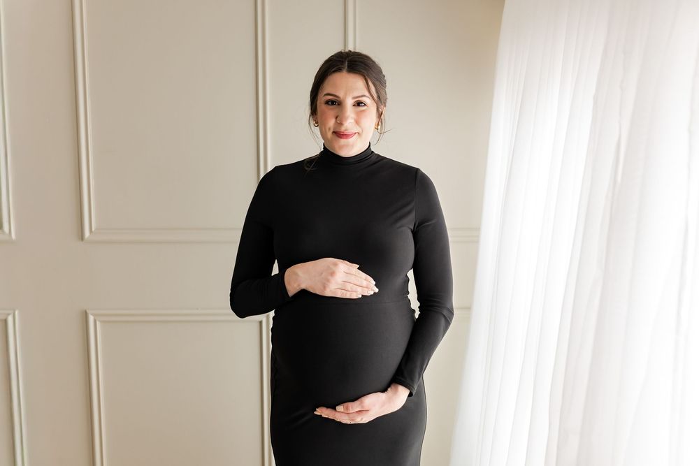 A pregnant woman in a black turtleneck dress stands in a bright white room showing her baby bump.