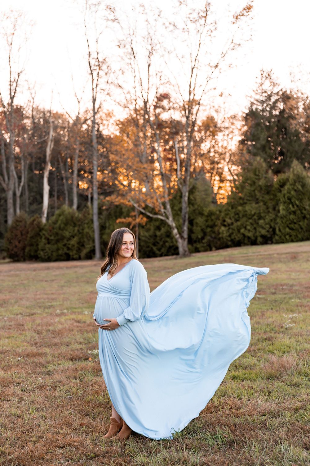 A flowing light blue dress billows in the wind during an outdoor maternity photoshoot at sunset.