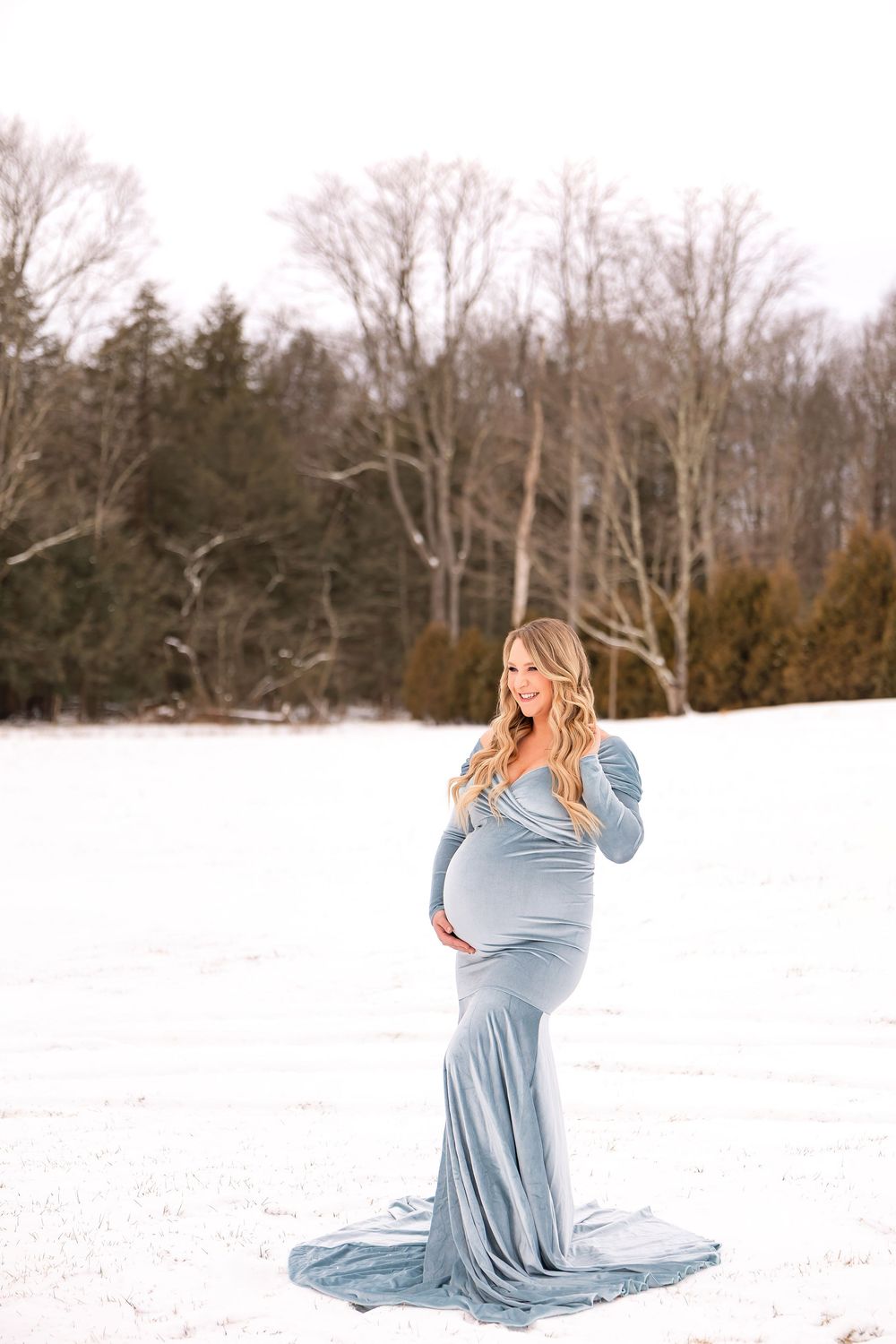 Woman in flowing light blue dress poses gracefully in winter snow scene with bare trees in background.