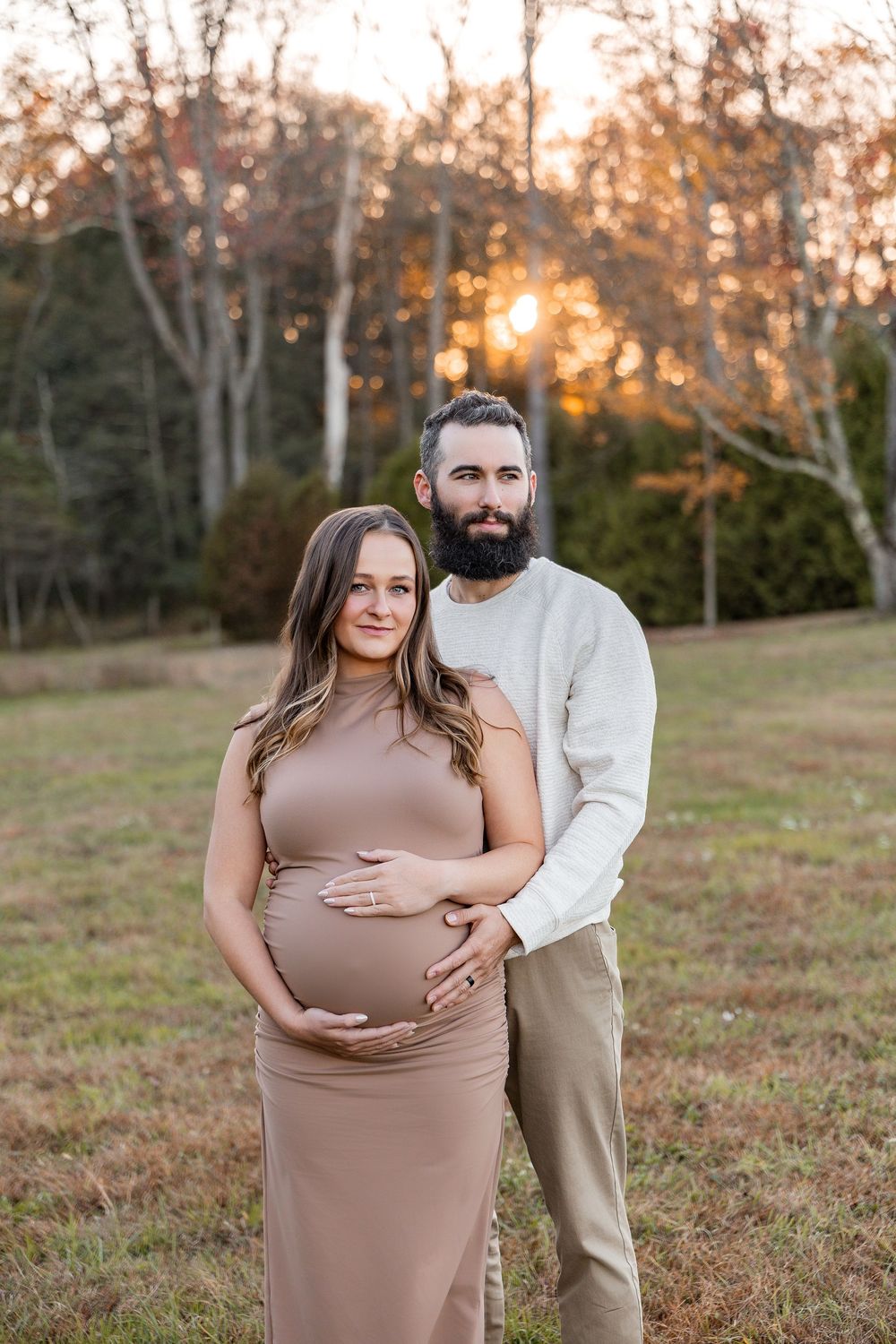 A couple poses together in a grassy field at sunset with autumn trees in the background.