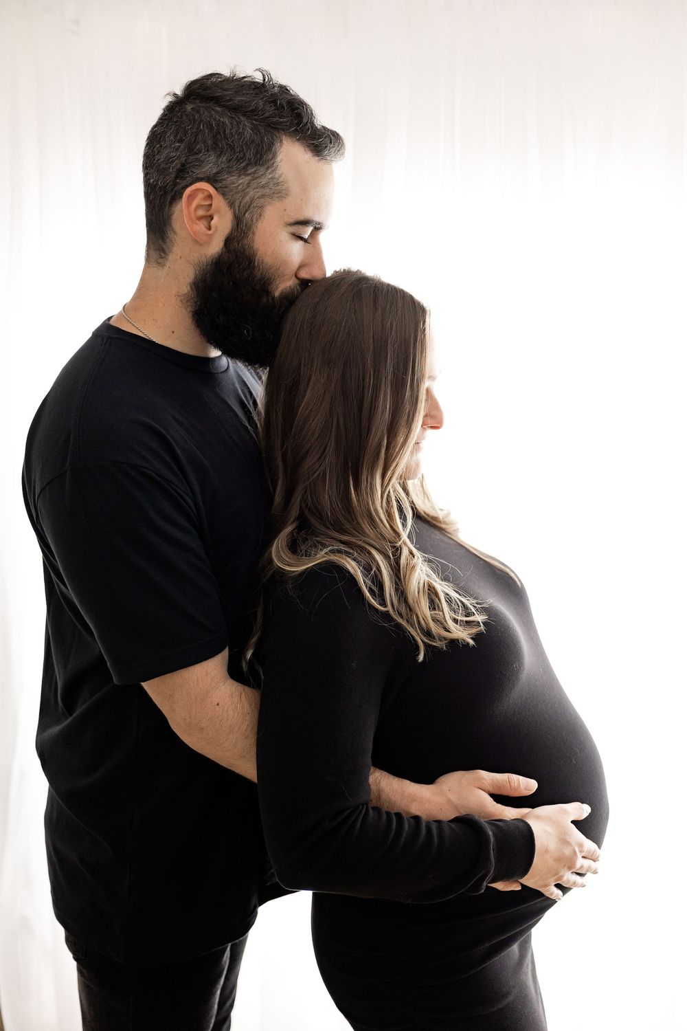A series of black and white maternity photos showing a couple in matching dark outfits embracing the pregnant belly.