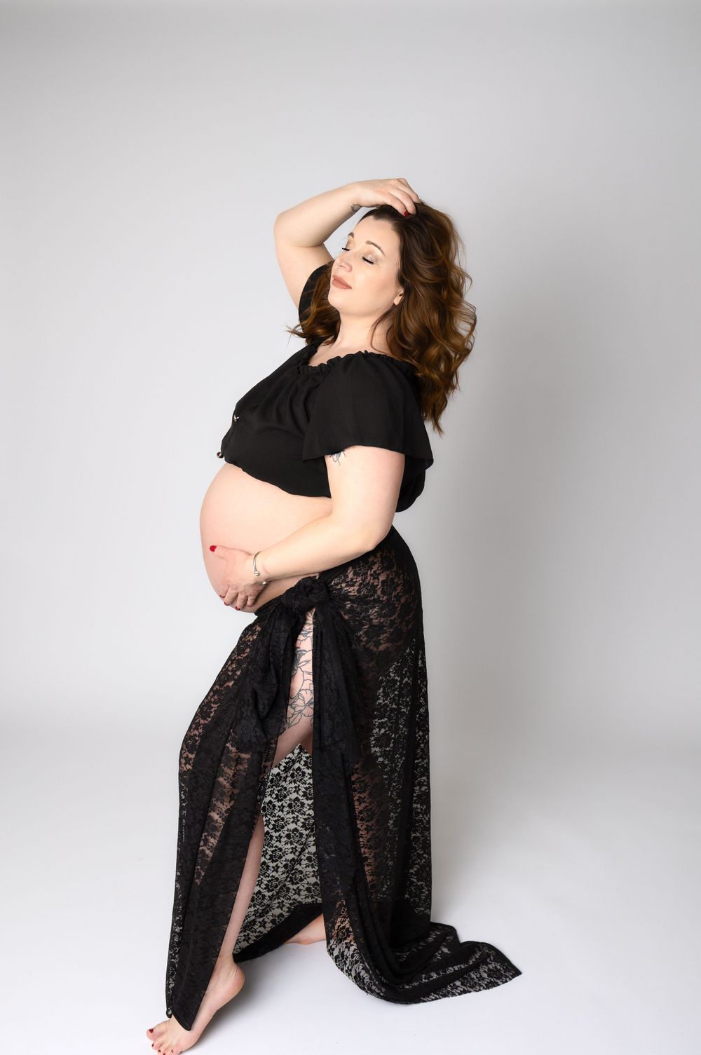 Artistic maternity pose in black lace dress against white background.