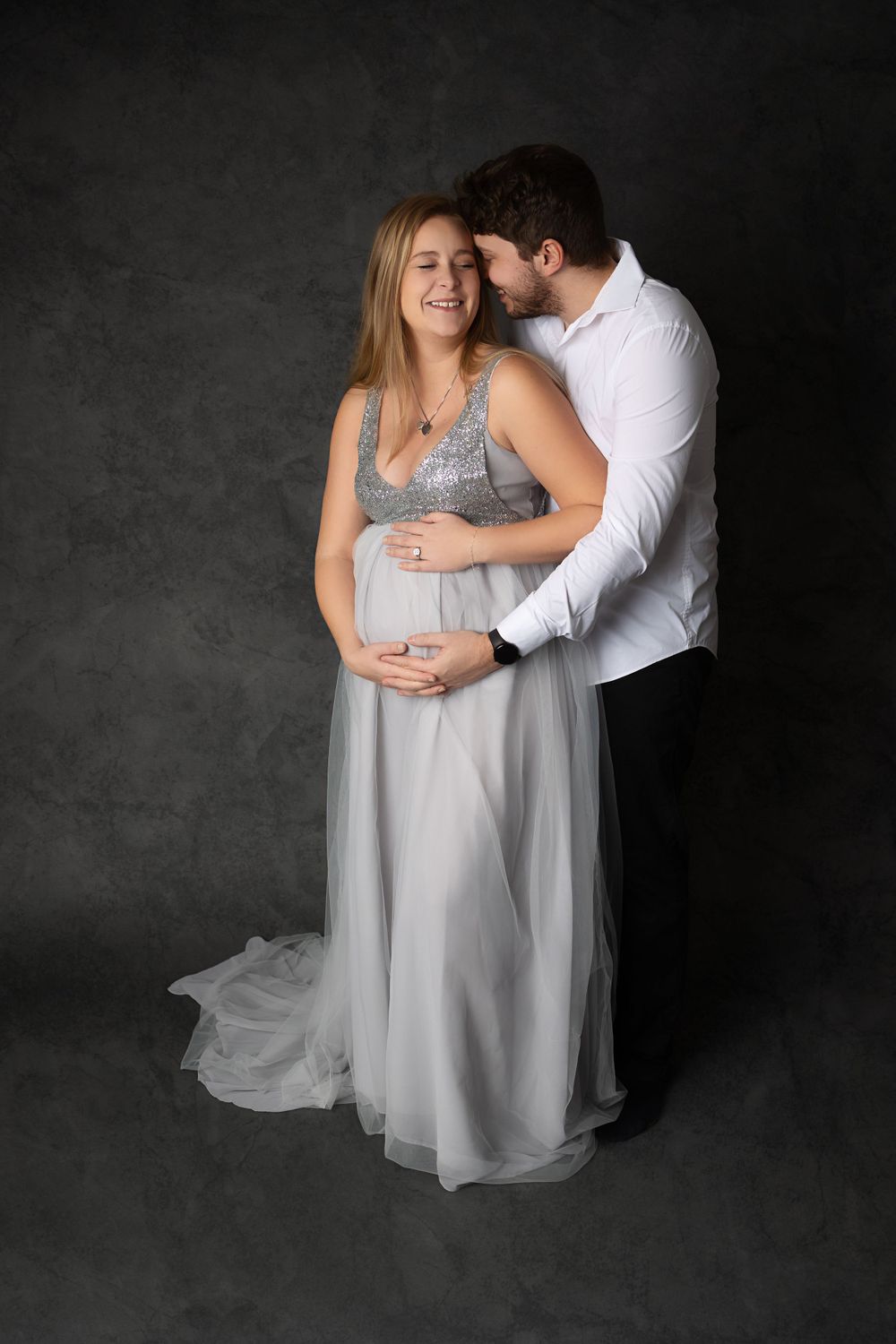 Intimate maternity portrait featuring couple in grey flowing dress and white shirt against dark background.