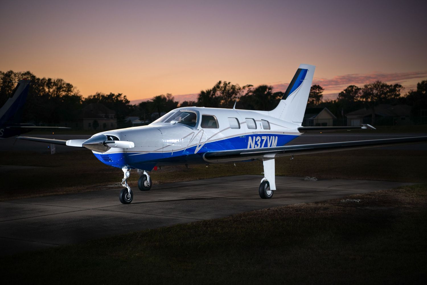 Small private aircraft parked on runway at sunset with dramatic orange sky and silhouetted trees in background.