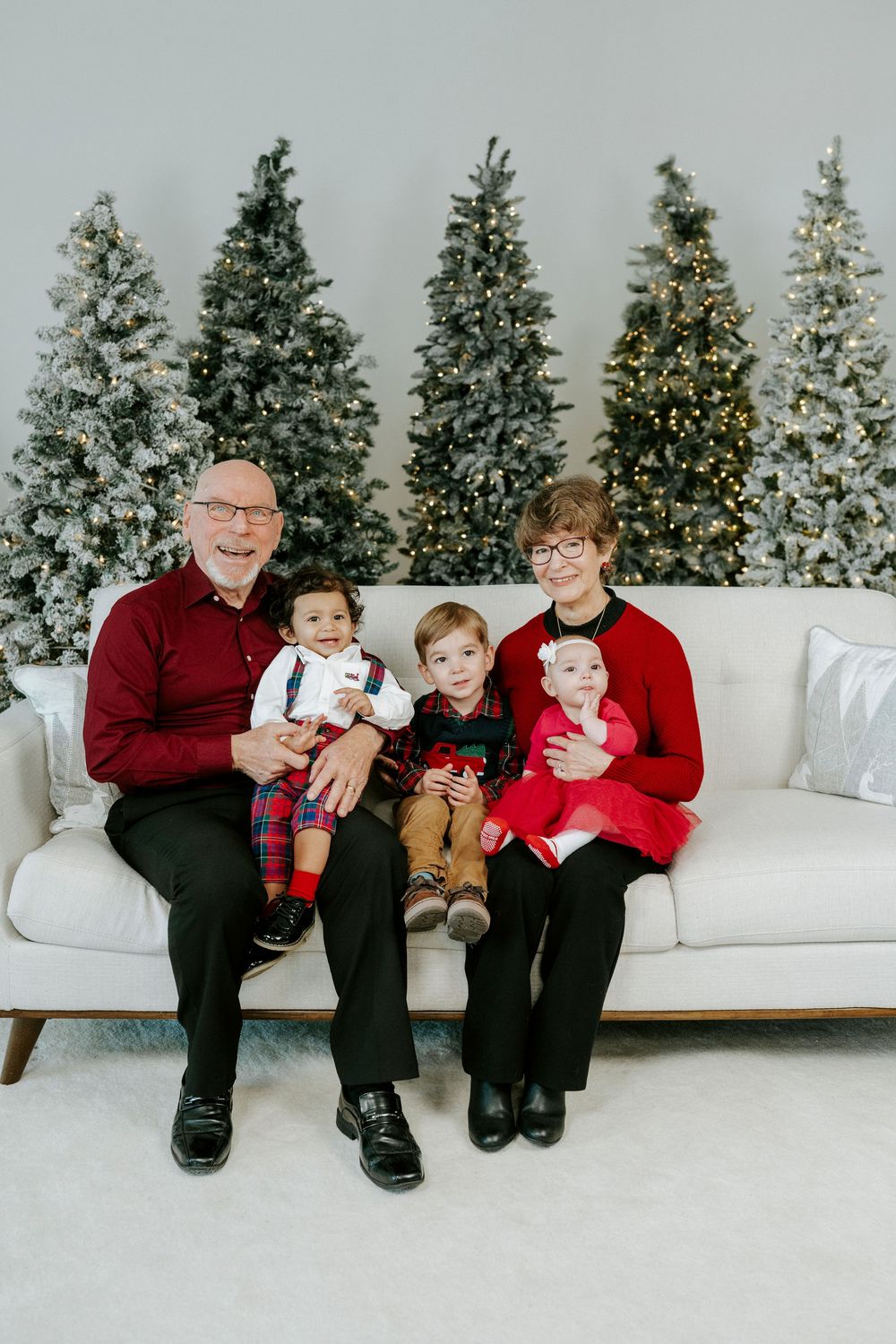 Four people in red and black holiday outfits sit together on a white couch with evergreen trees in the background.