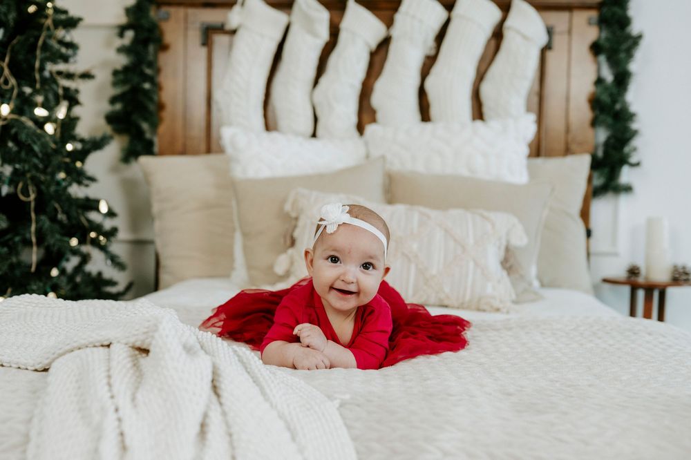 Baby in red blanket lying on white bedding with holiday stockings and greenery decorations on wooden headboard.