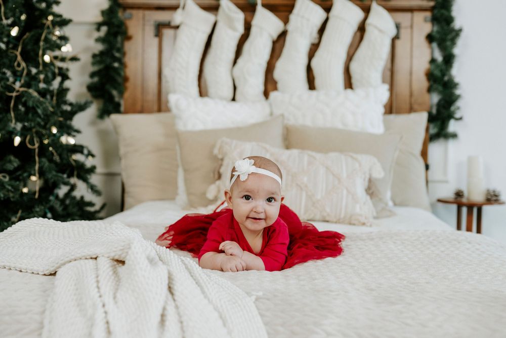 A baby wearing a red sweater lies on white bedding while playing and smiling in a cozy bedroom setting.