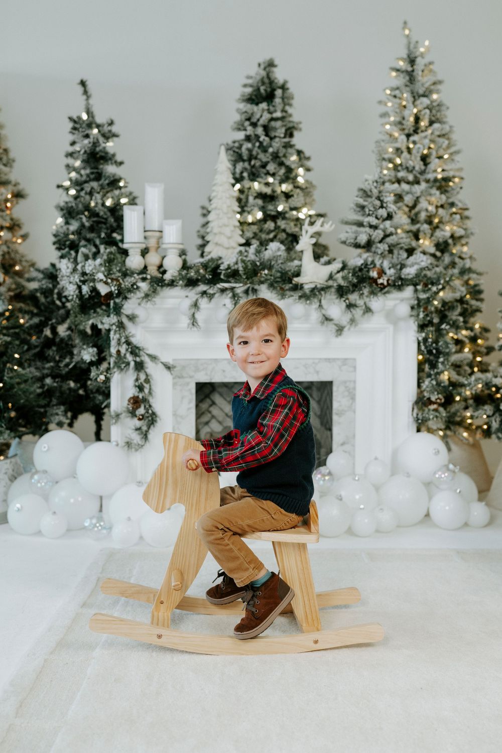 Holiday scene with decorated Christmas trees, white fireplace, and wooden rocking horse against snowy white backdrop.
