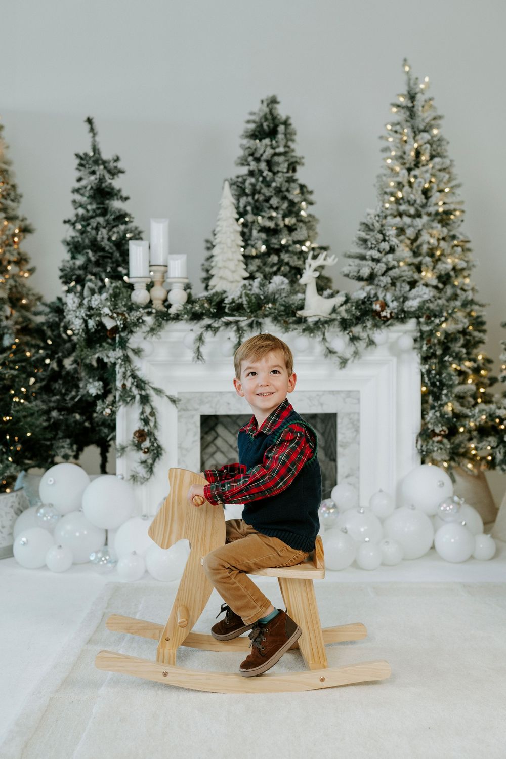 Holiday scene with decorated Christmas trees, white fireplace, and wooden rocking horse surrounded by white balloons.