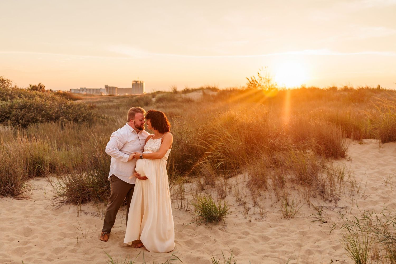 A couple embraces on a sandy beach during golden hour sunset with tall grass and dunes in the background.