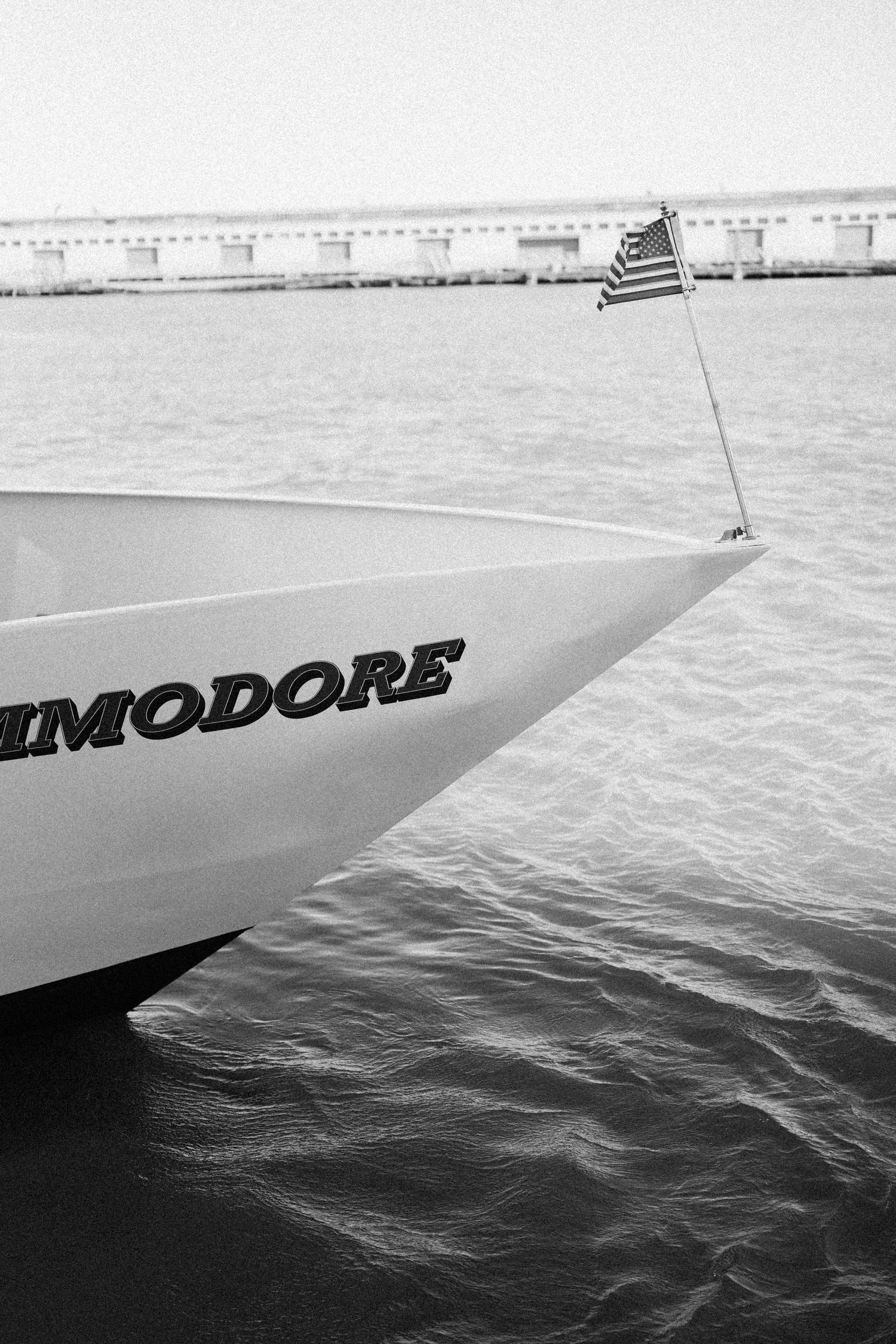 Black and white view of a yacht's bow cutting through water with an American flag visible.