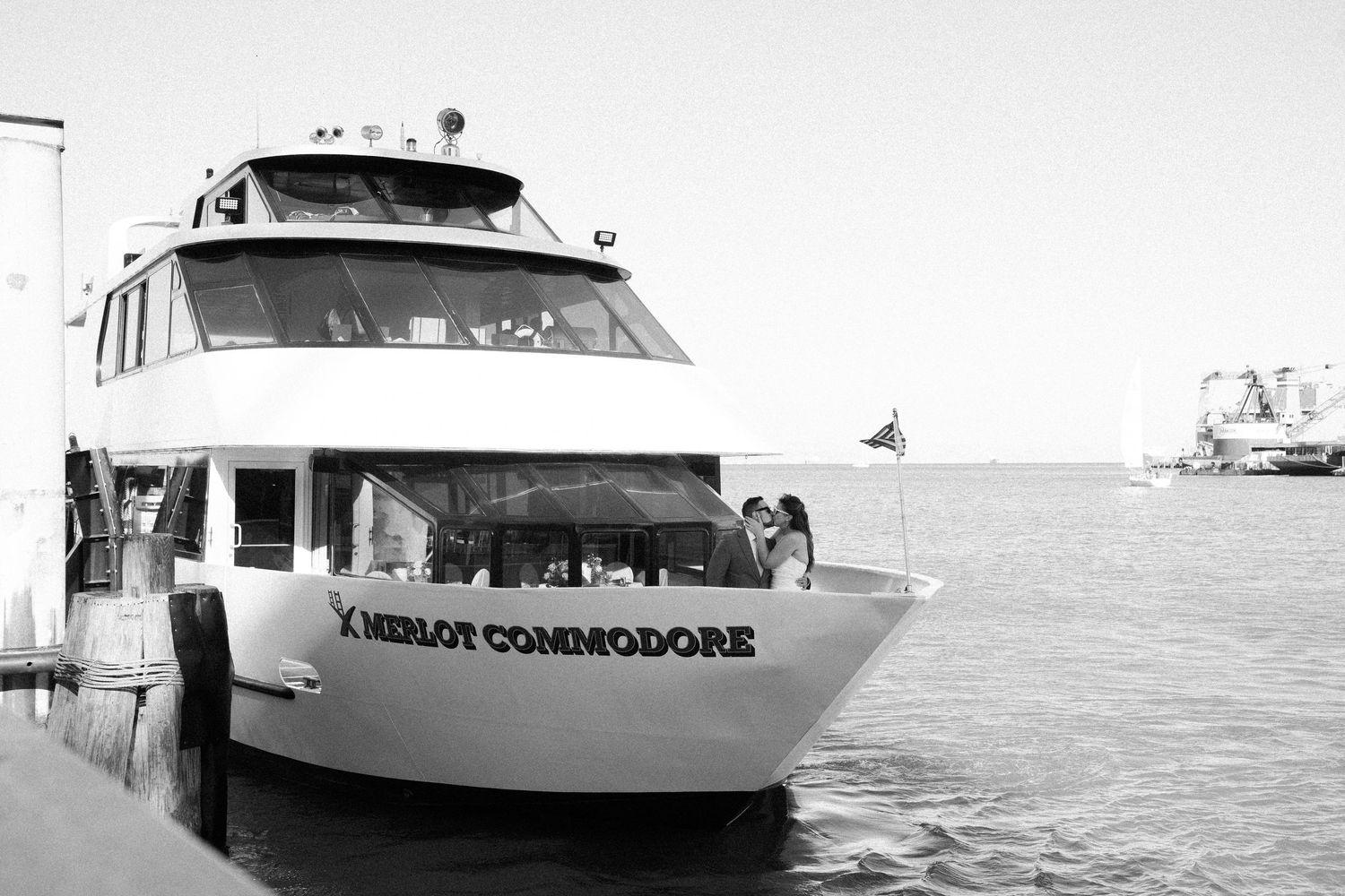 A black and white photograph of a yacht named Commodore docked at a marina with water and boats in the background.
