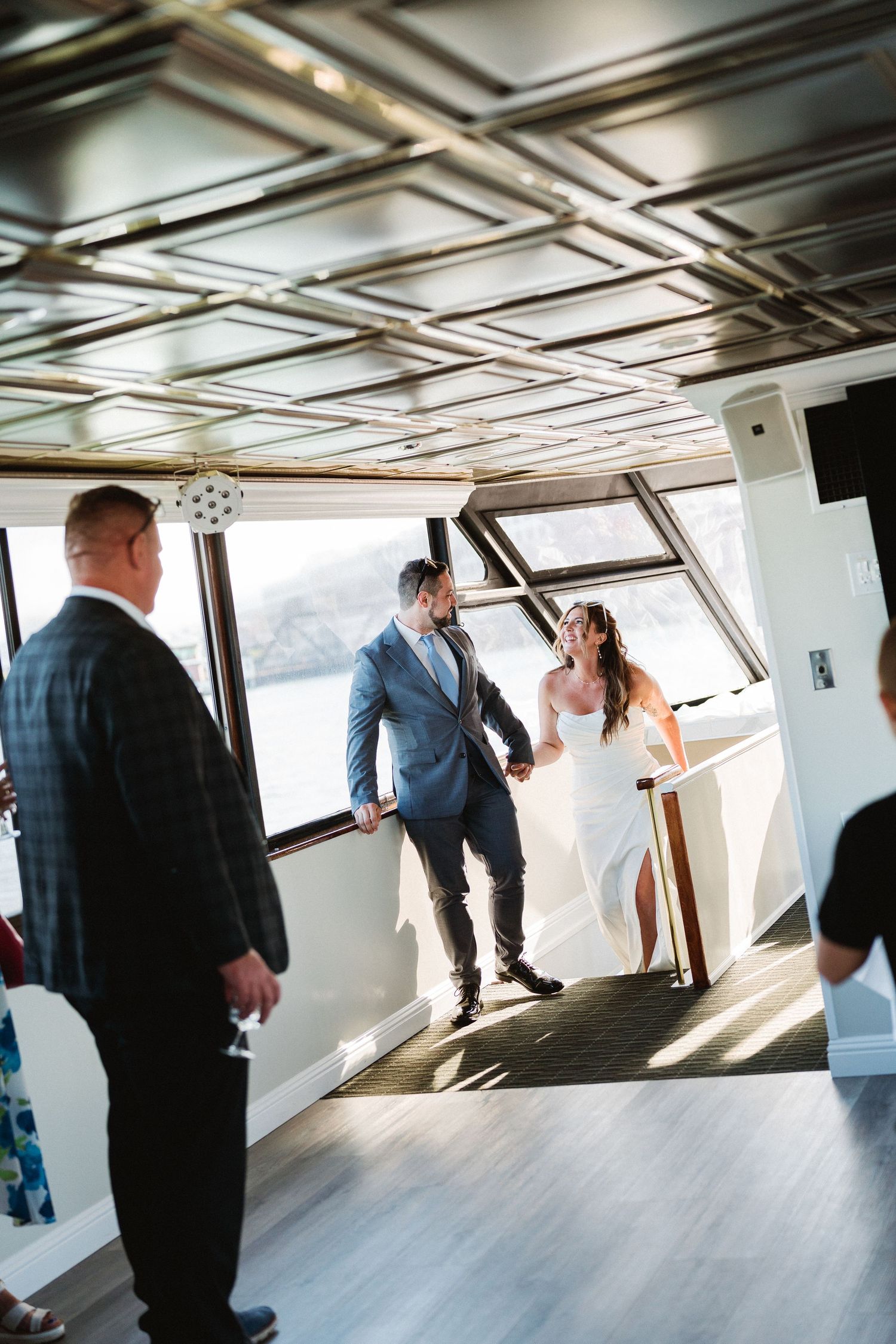 Silhouettes of wedding guests mingling in a sunlit yacht interior with geometric ceiling patterns.