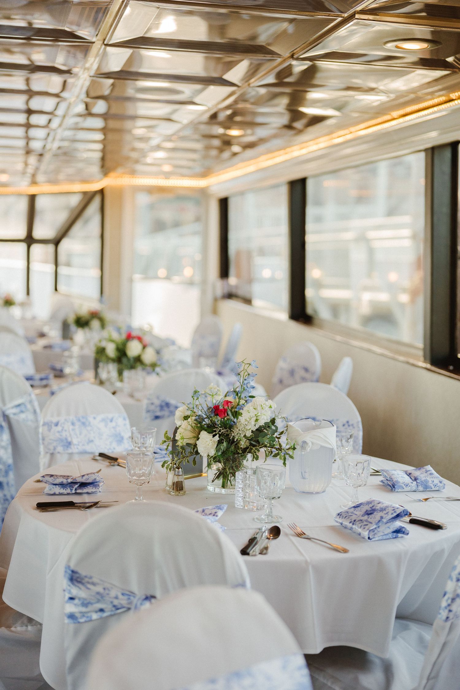 Elegant wedding reception tables set with white linens, blue accents and floral centerpieces in a bright indoor venue.