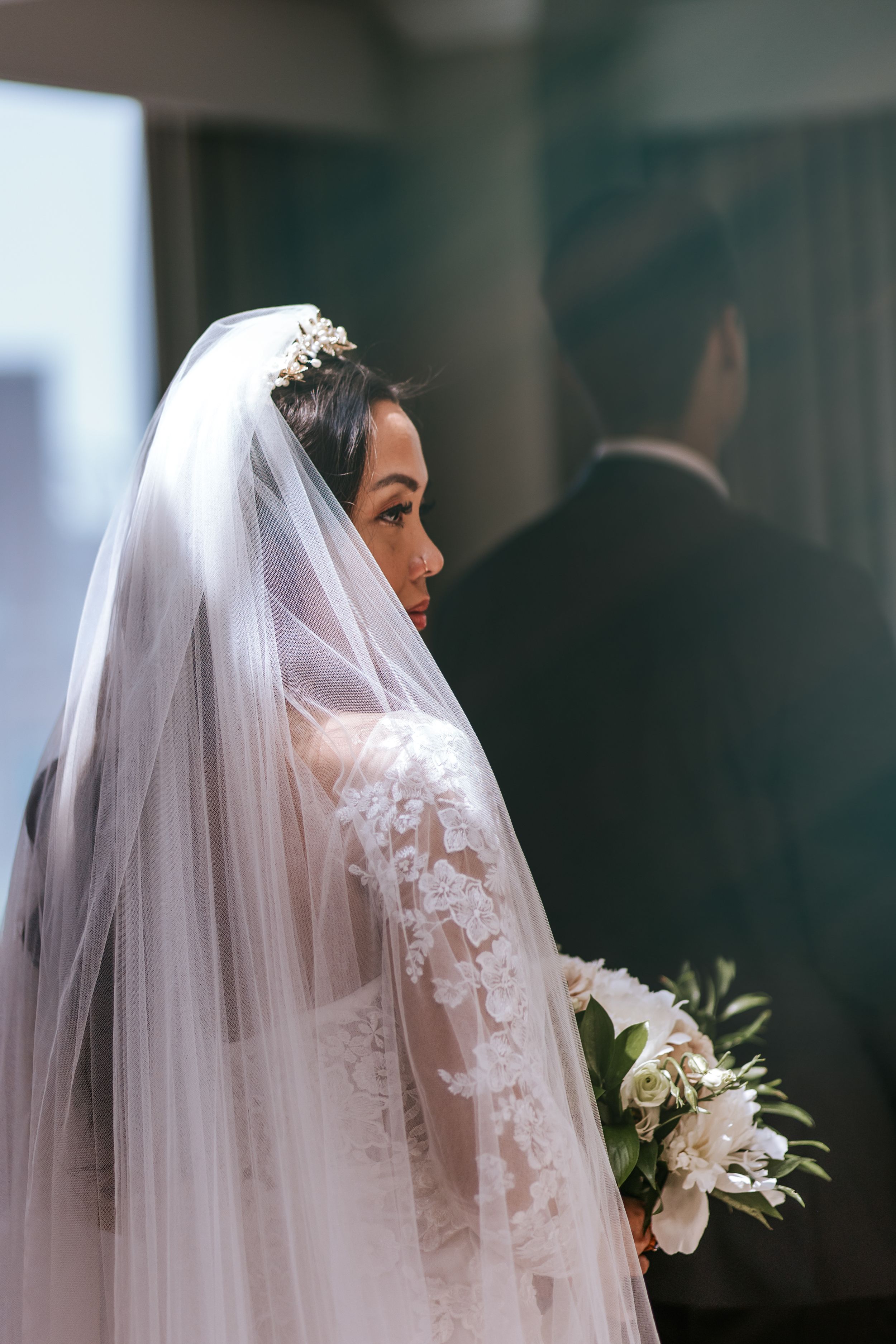 A bride in a white lace wedding dress and veil stands in profile against a dark background.