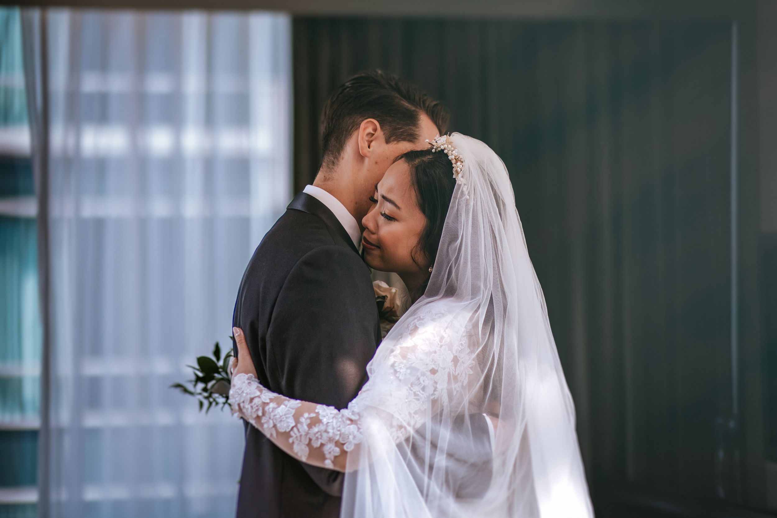 A romantic wedding couple shares an intimate moment in front of a window with soft natural lighting.