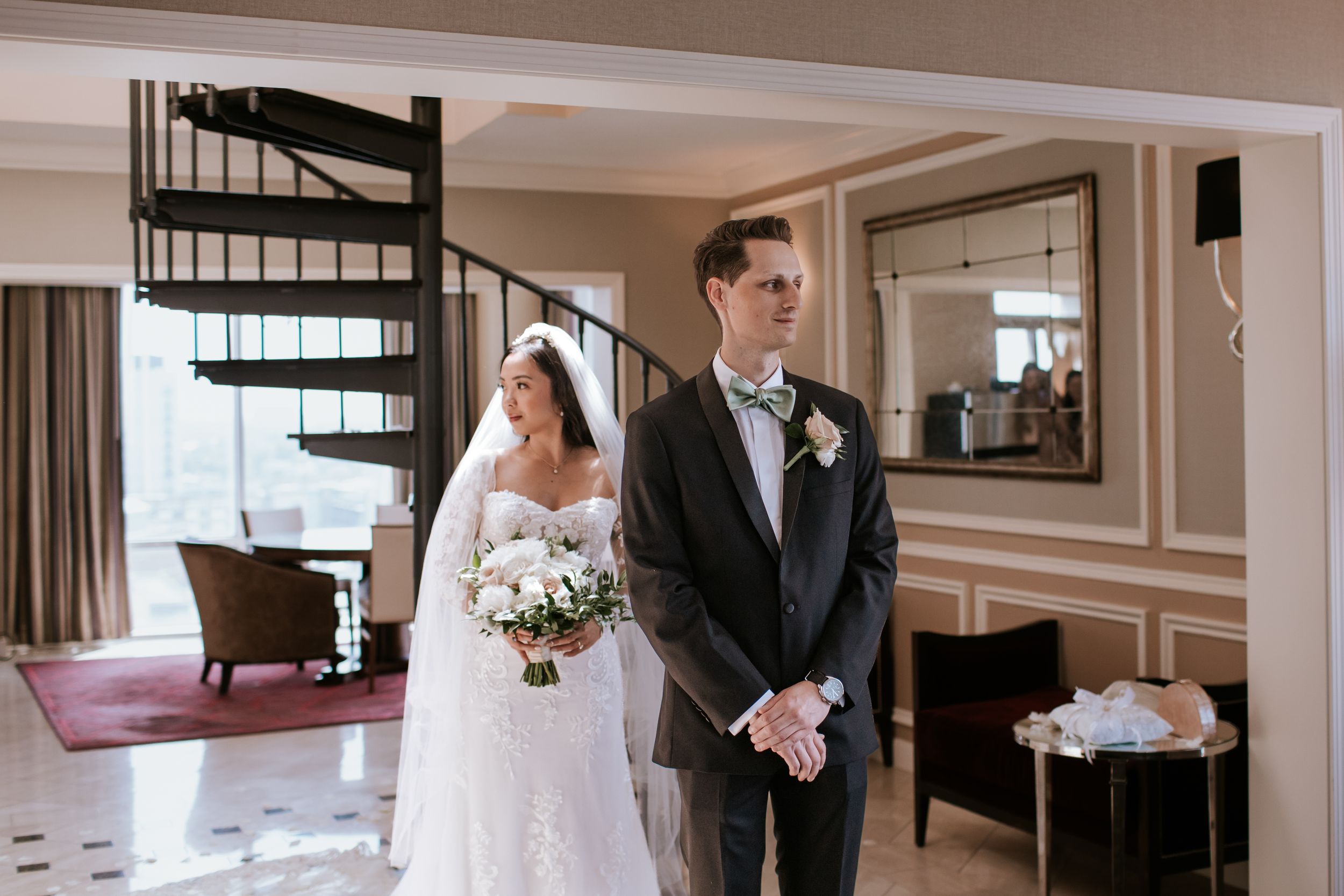 A wedding party poses in an elegant indoor venue with cream colored walls and a spiral staircase in view.