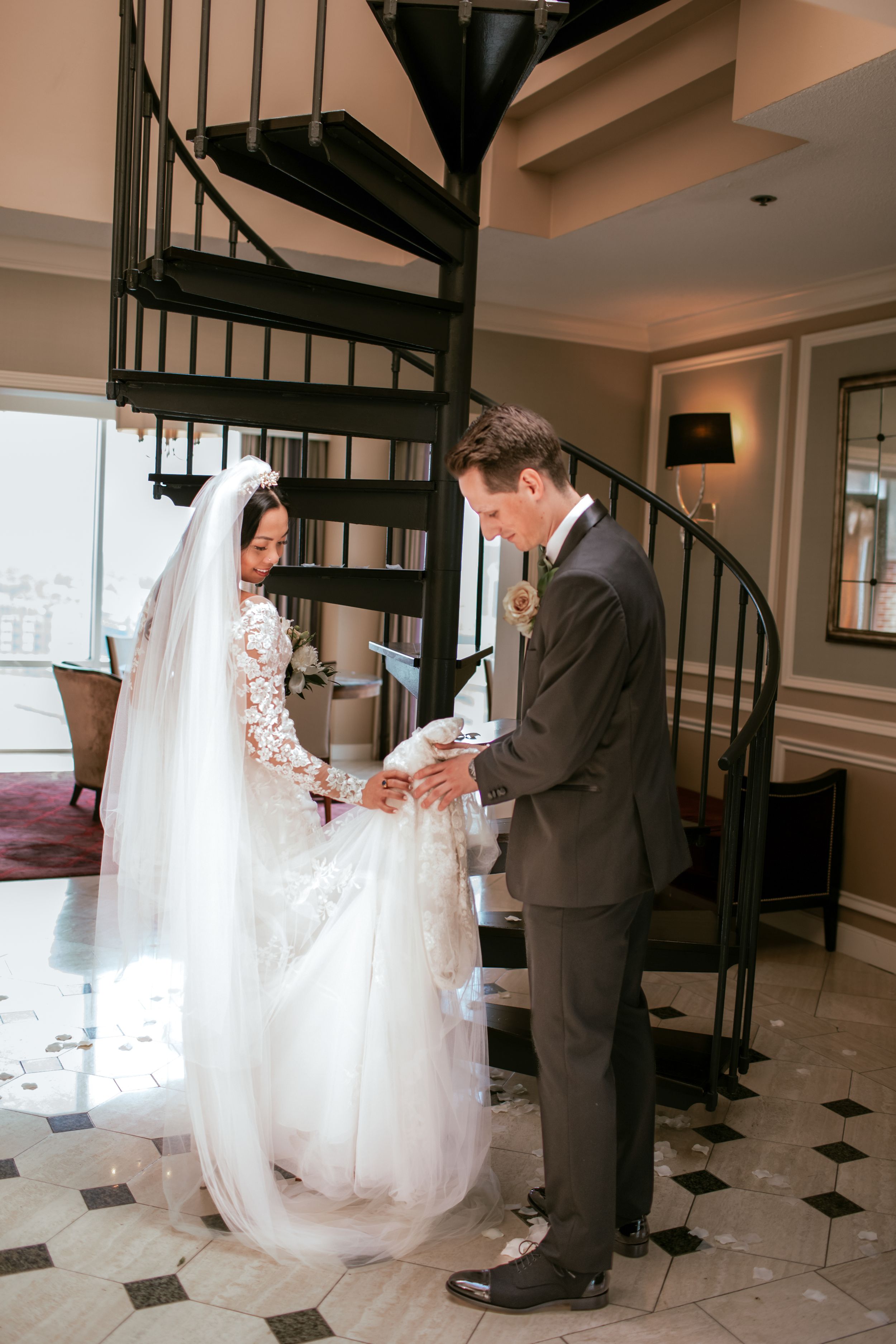 A wedding couple stands at the bottom of a spiral staircase in a black and white tiled room during their first look moment.