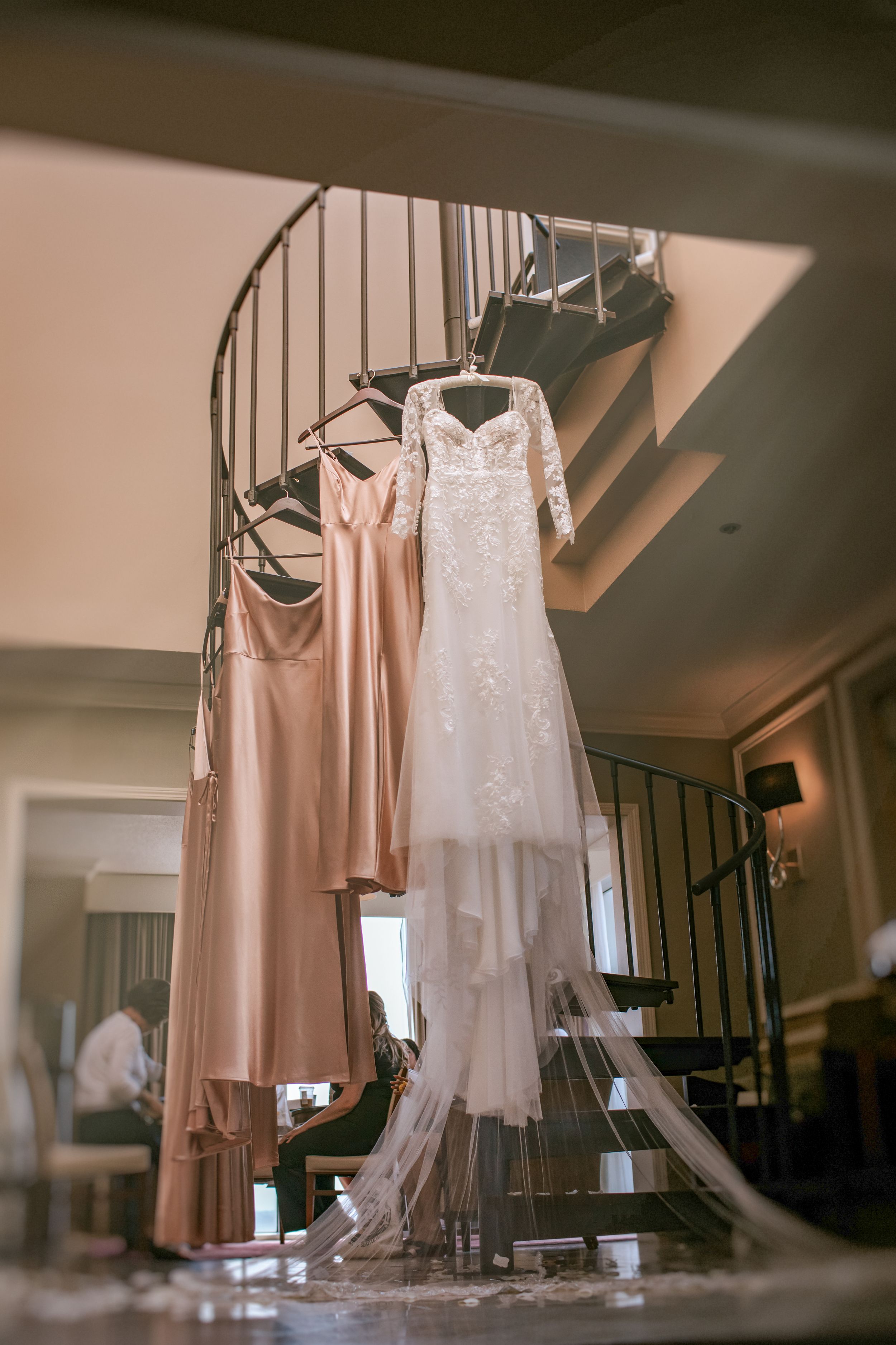 Elegant wedding dresses and bridesmaid gowns in blush pink and white hang from hangers in a dimly lit room.