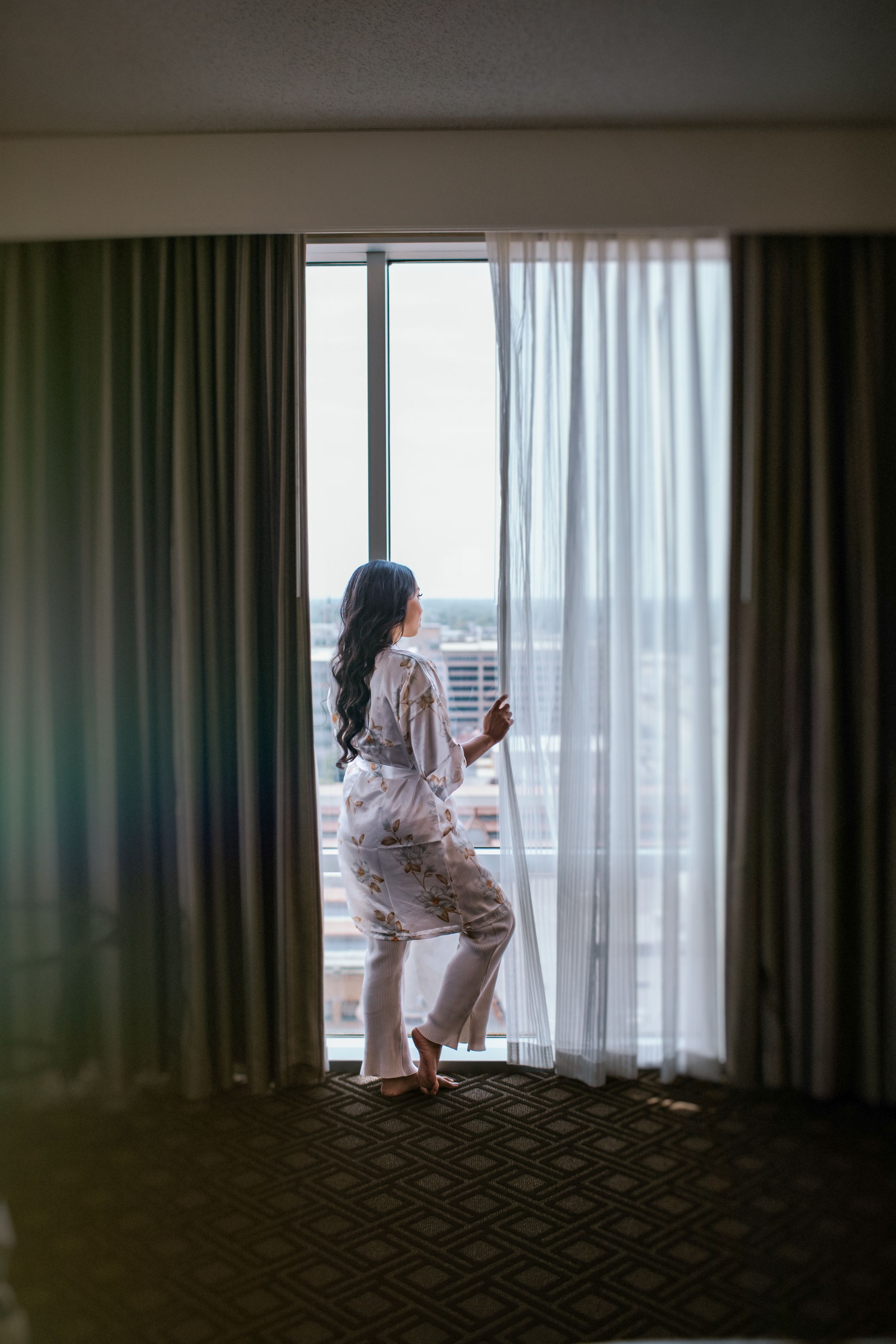 A person in sleepwear stands by floor-to-ceiling windows with sheer curtains, looking out at a city view.