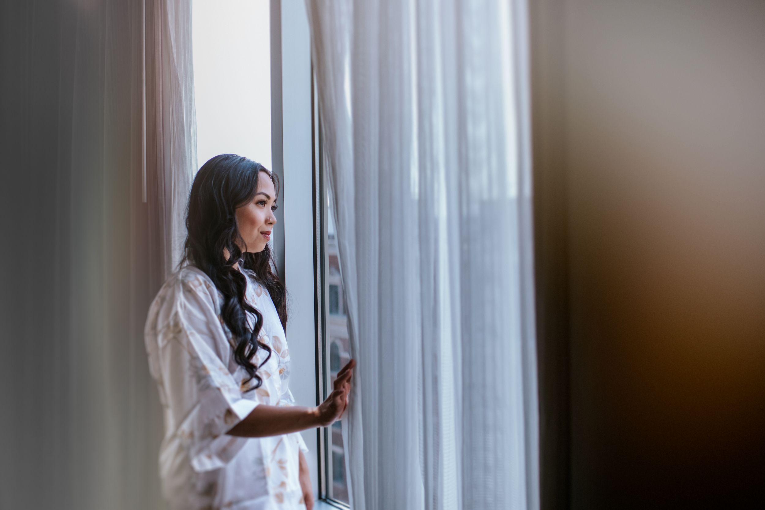 Woman in white sweater stands by window while looking at smartphone in hand.