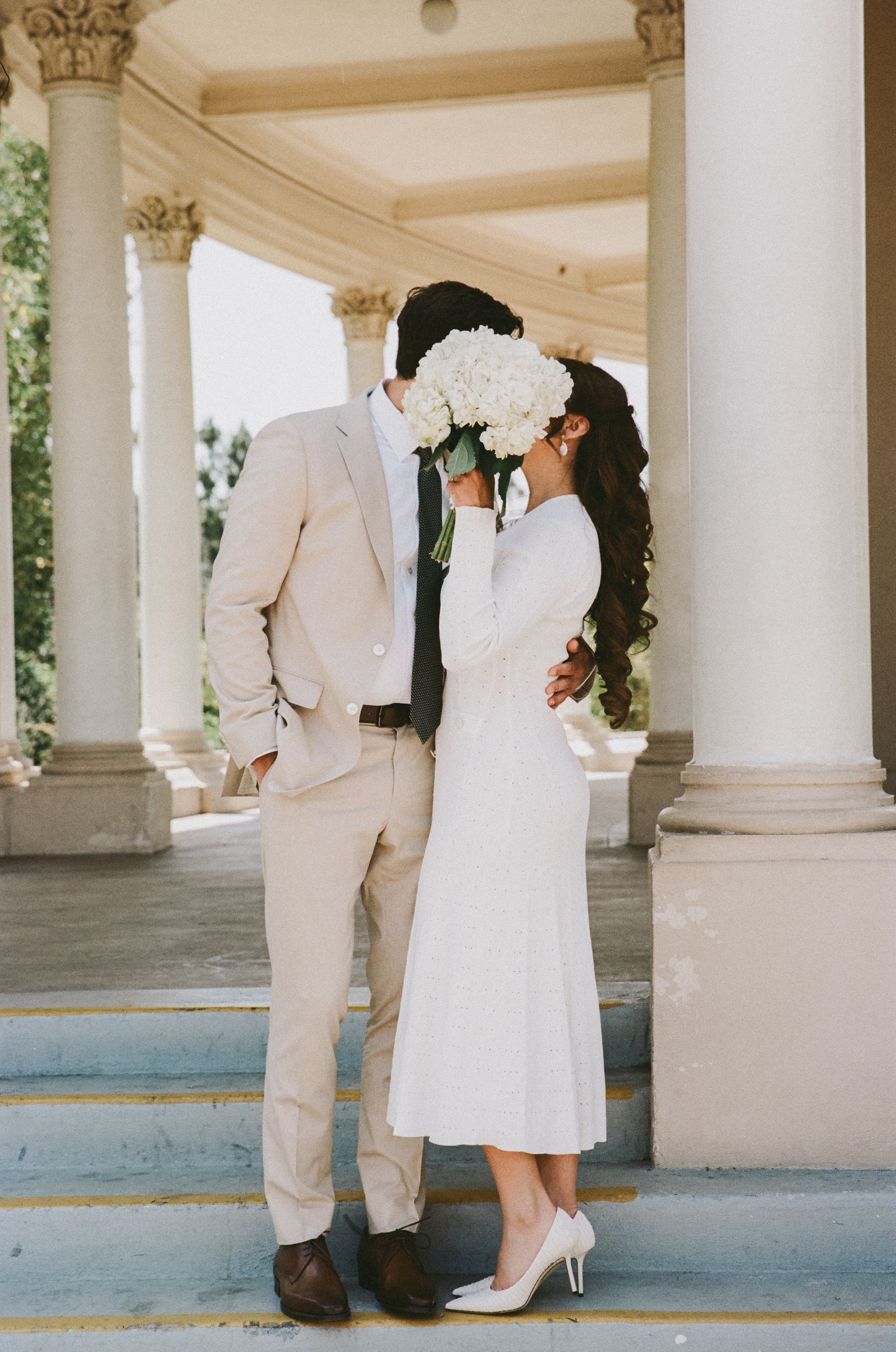Couple in white attire poses beneath classical white columns of an ornate pavilion.