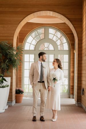 Couple in light-colored formal wear poses in an elegant room with arched windows and terracotta floors.