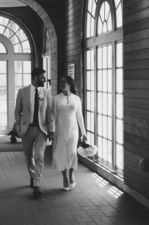 Black and white photo of a couple walking through a sunlit hallway with arched windows.