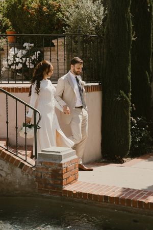 Couple in white outfits walks along a brick wall path in a garden setting.