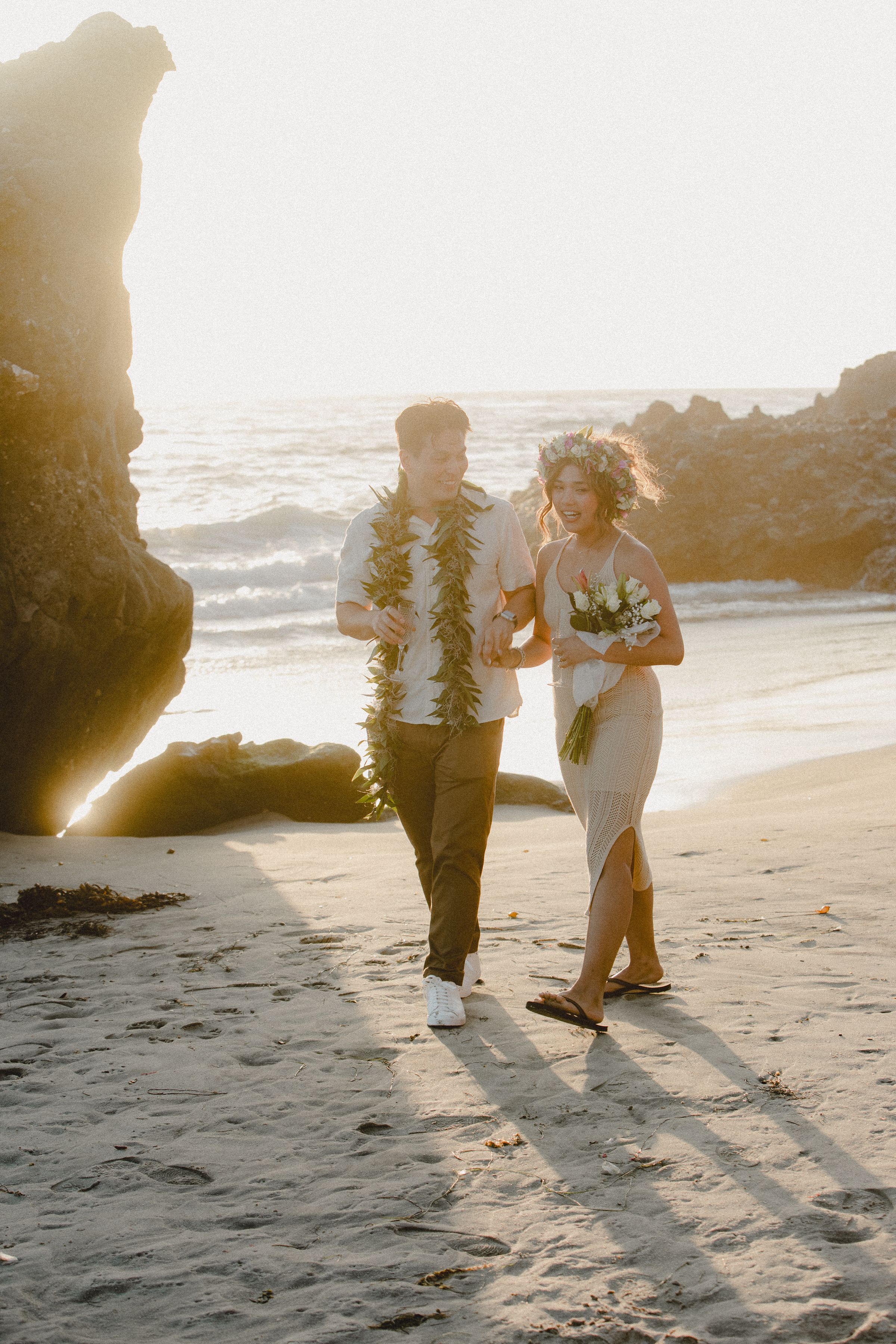 A couple wearing leis walks along a beach at sunset with dramatic rock formations in the background.
