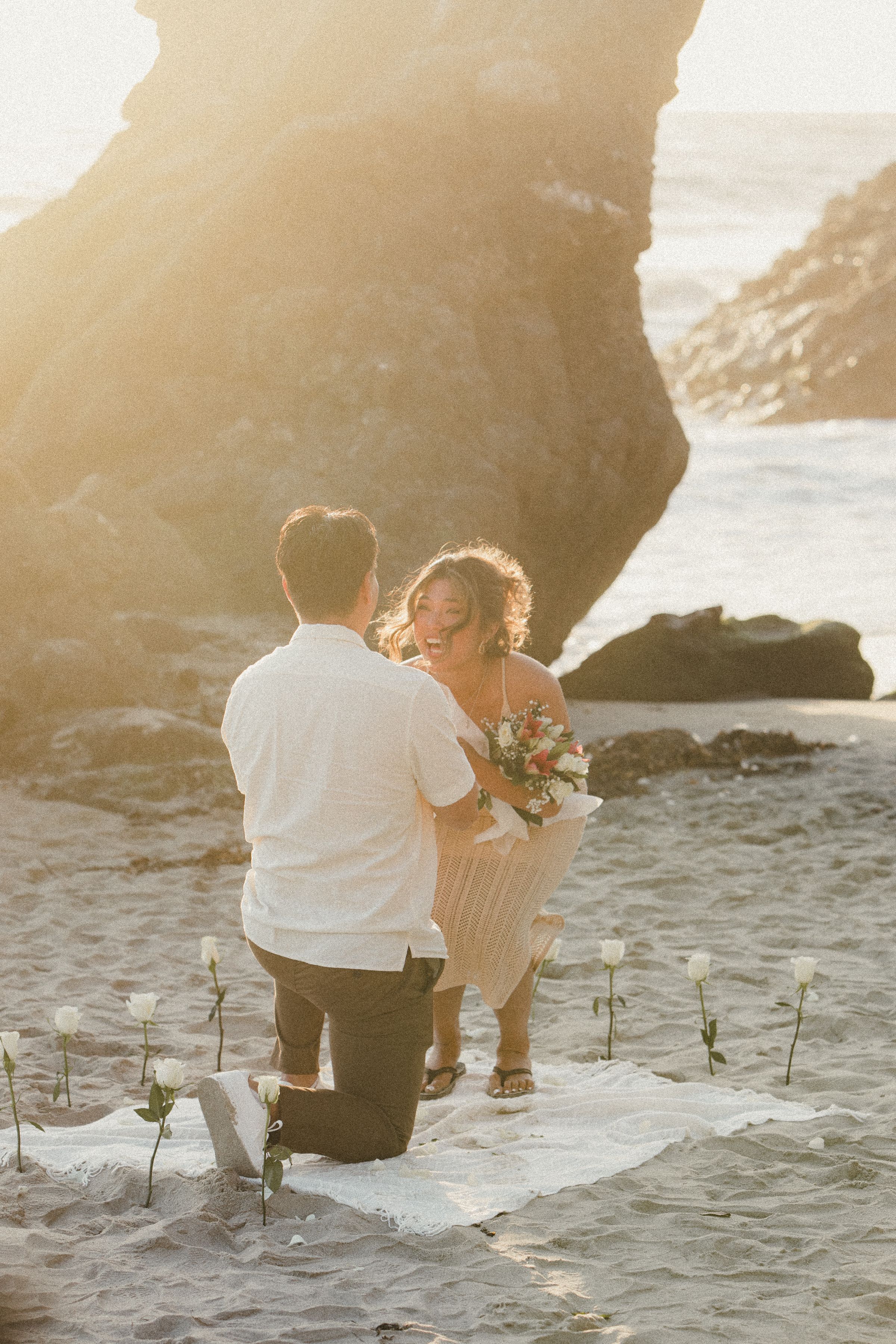 A romantic beach proposal scene with crashing waves and rocky cliffs at sunset.