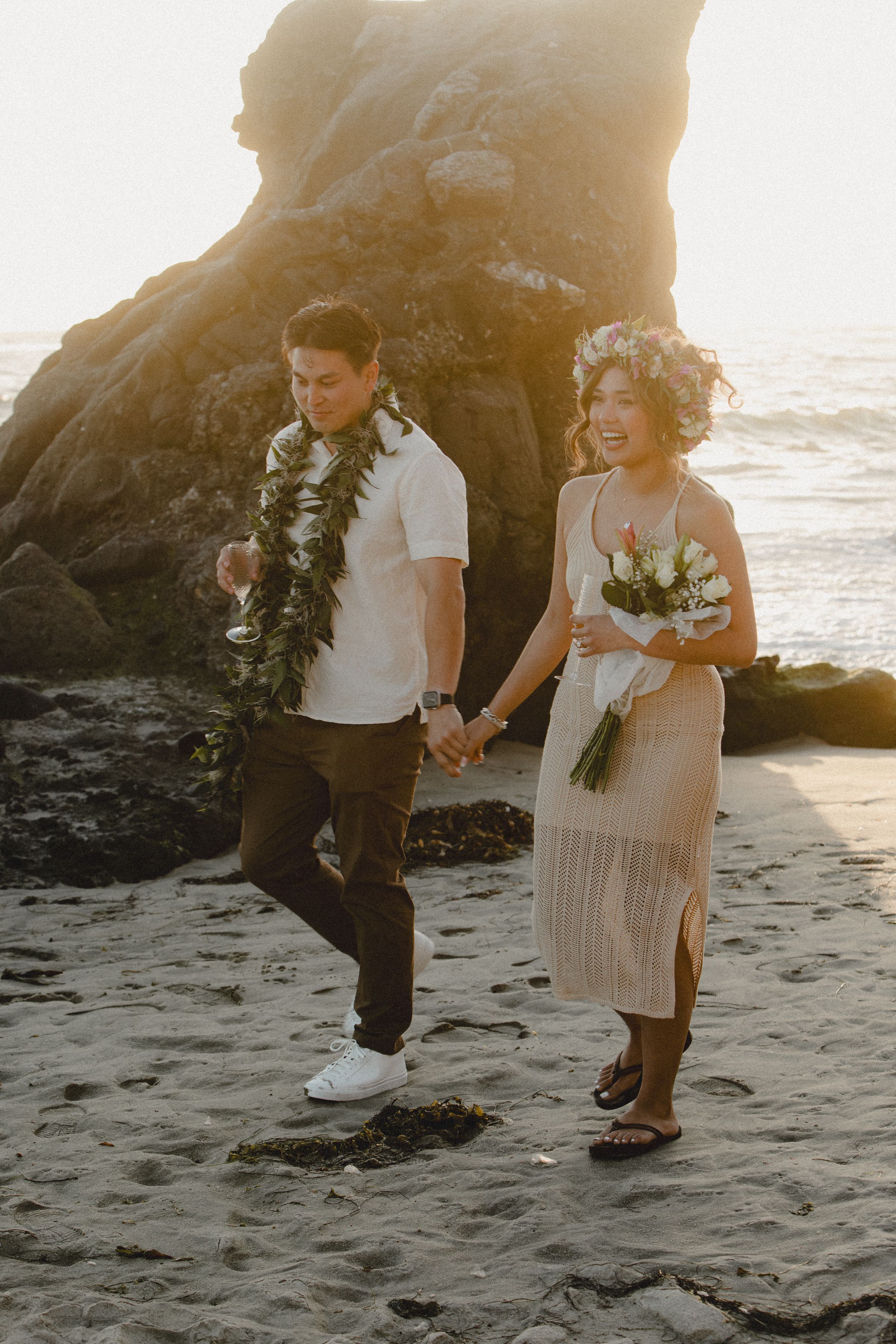 A couple in beach wedding attire walking hand in hand along the shore at sunset.