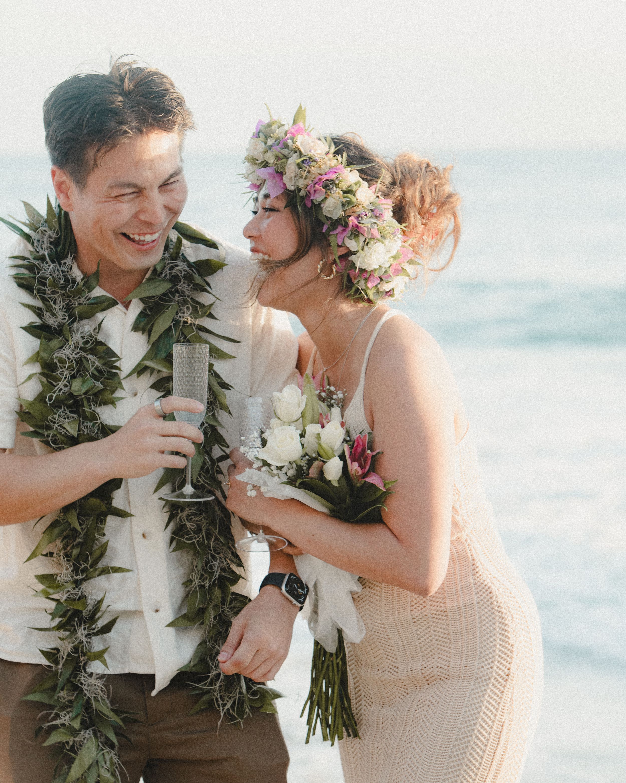 A beachside couple wearing floral leis and wedding attire share a joyful moment by the ocean.