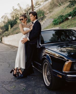 Romantic couple shares intimate moments beside a classic black car on a scenic hillside road.
