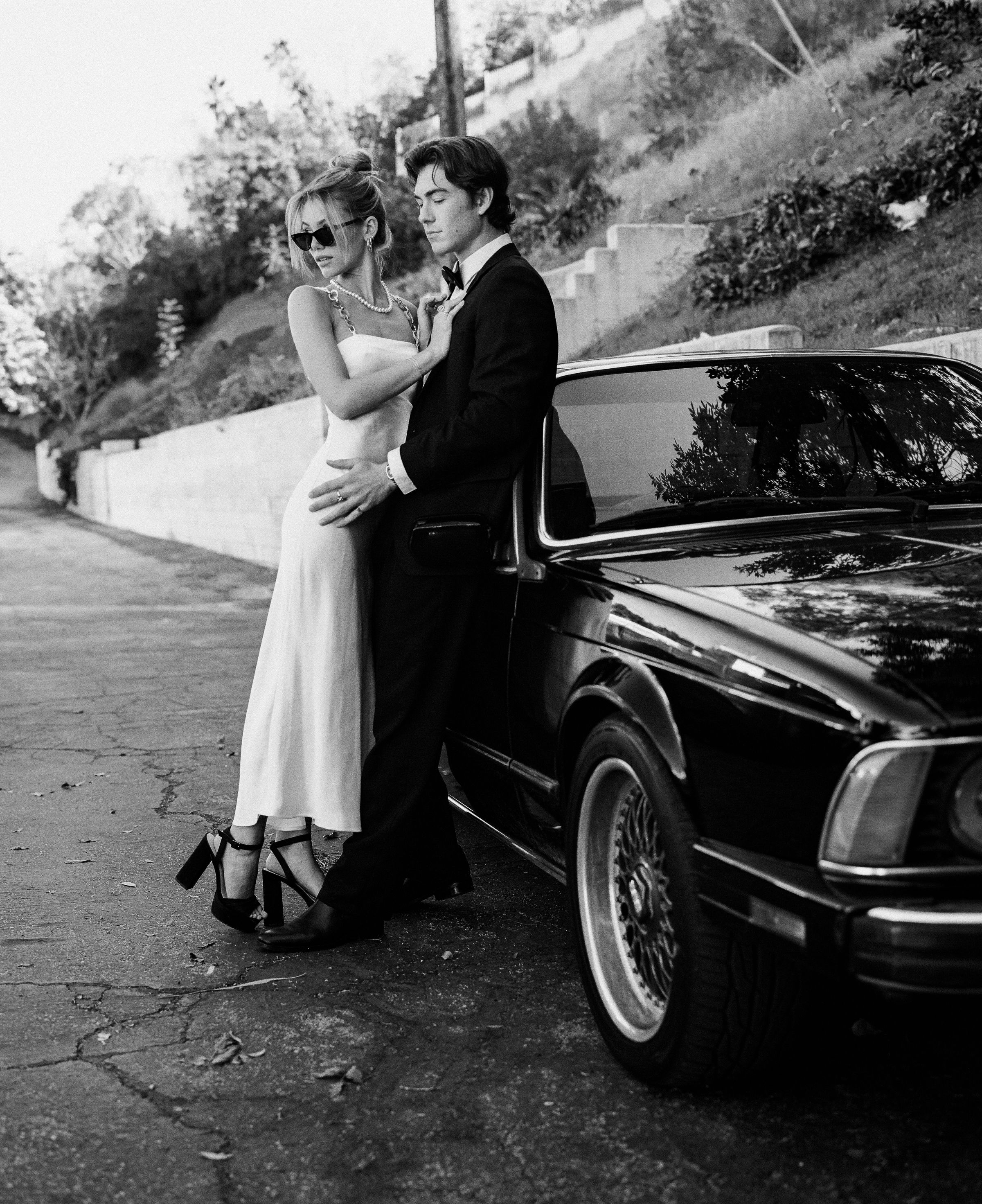 A vintage-style photo of a couple in formal attire posing beside a classic car.