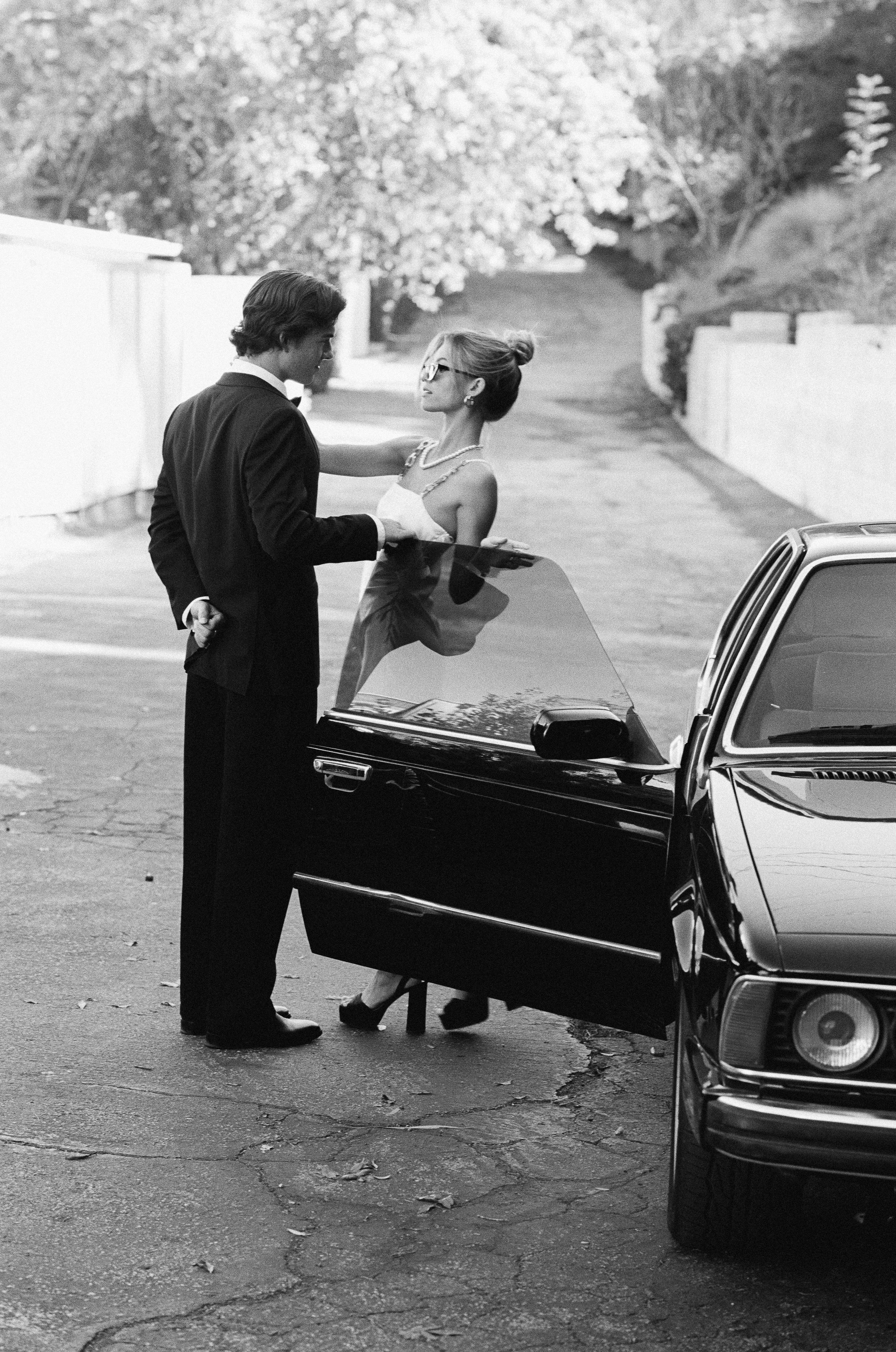 A vintage black and white photo of a couple in formal wear beside a luxury car.
