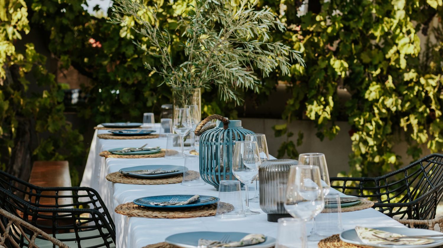Elegant outdoor dining table set with blue plates, wicker chargers, crystal glasses, and a lush floral centerpiece in sunlight.