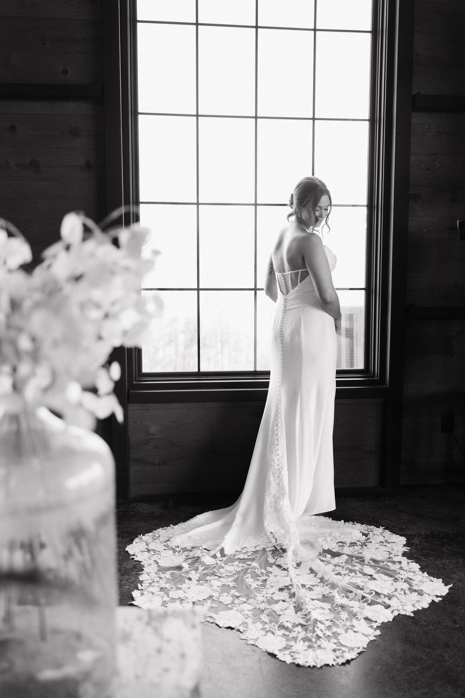 A bride in a flowing white lace gown poses gracefully against a window in a rustic wooden room.