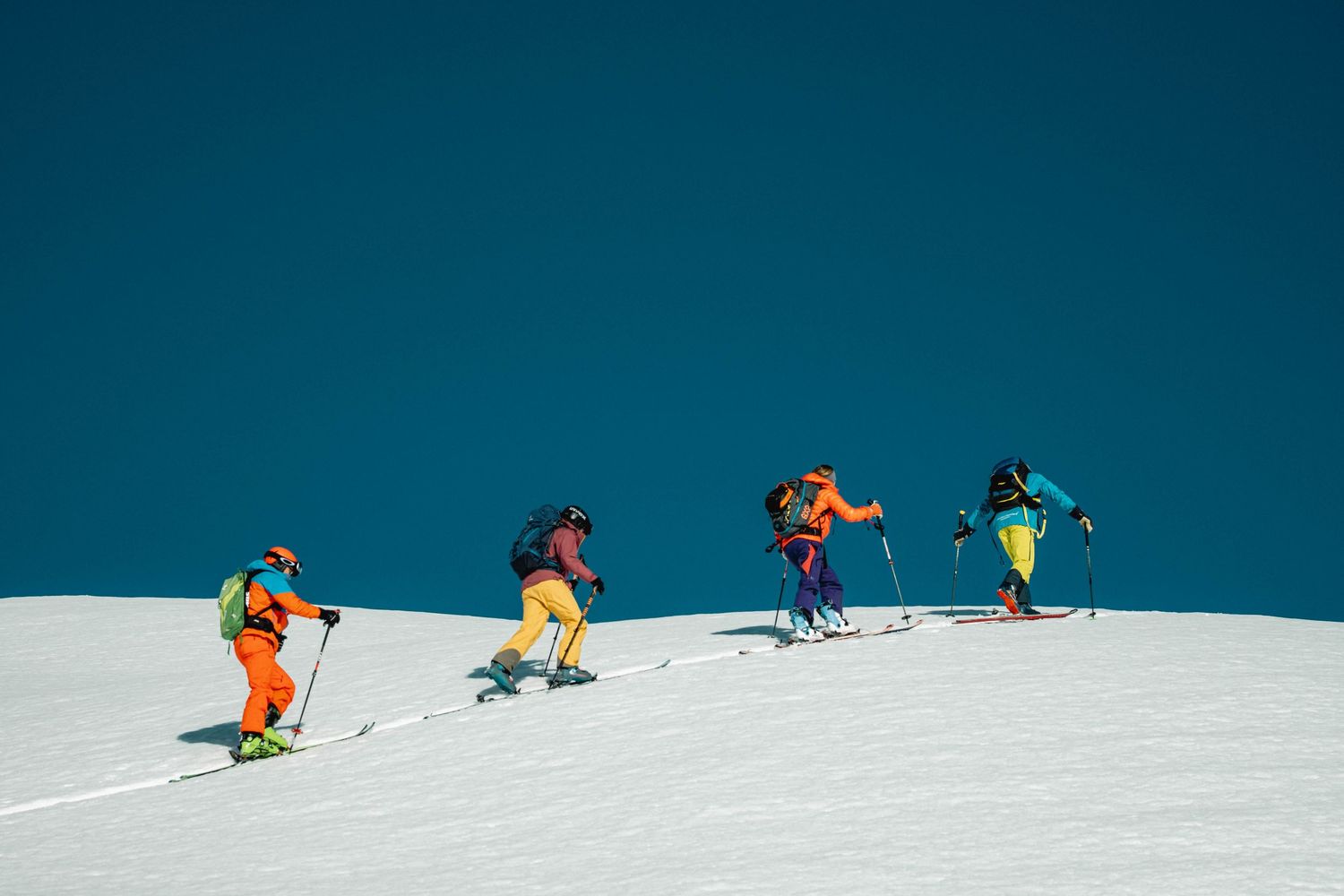 Four skiers in bright winter gear climb up a snowy slope against a vibrant blue sky on a sunny day.