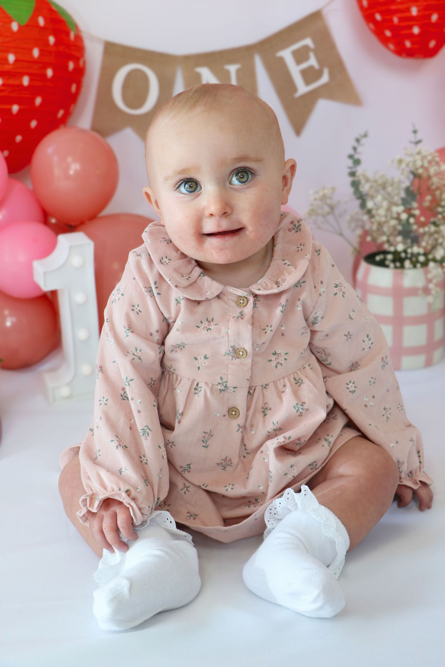 A baby in a pink floral dress and white booties sits against a backdrop with strawberry decorations and pink balloons.