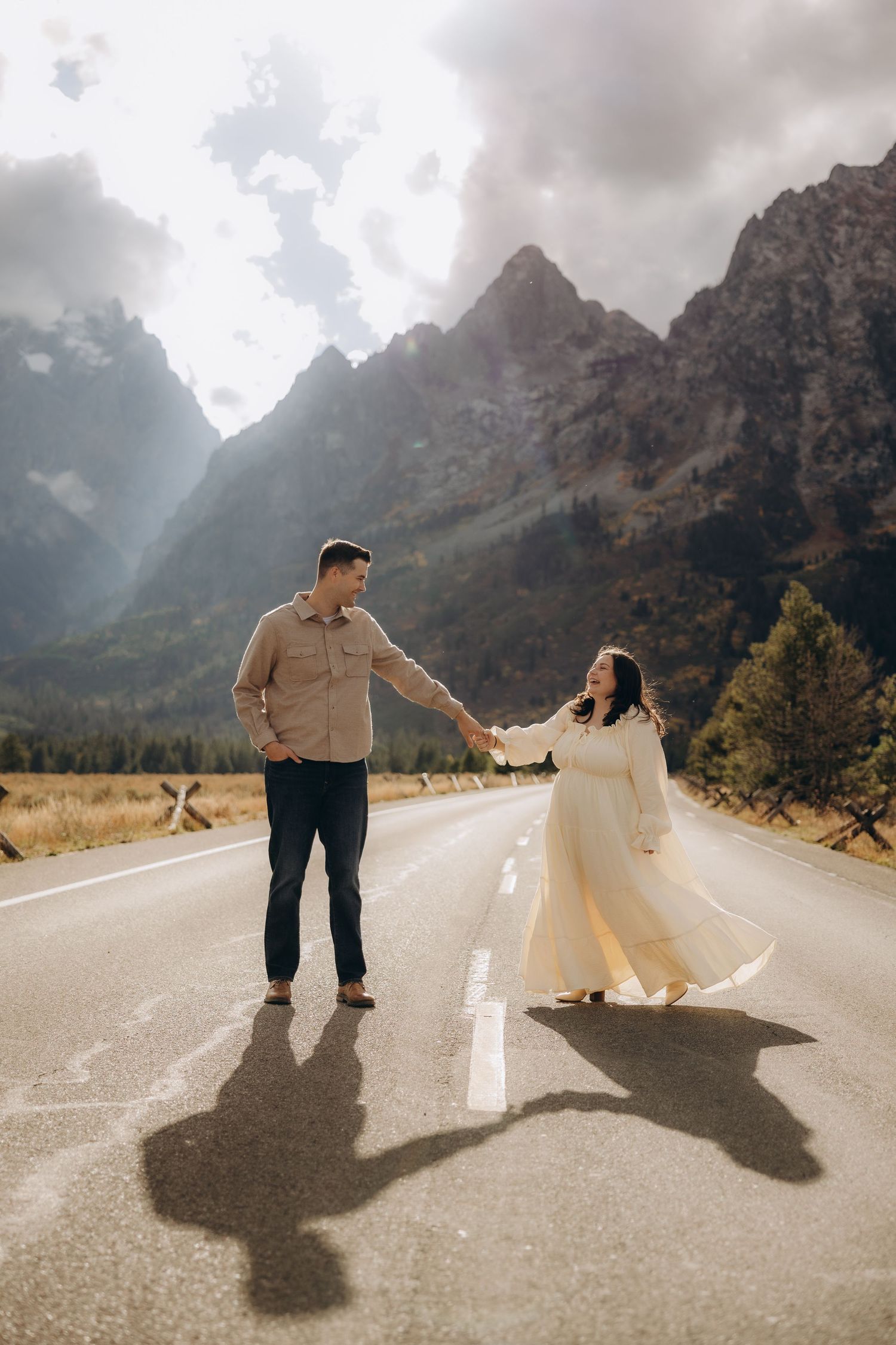 A couple dances together on a mountain road with dramatic peaks and cloudy skies in the background.