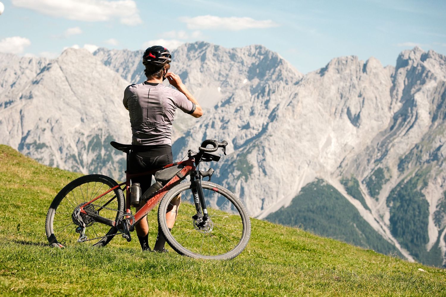 A cyclist takes a break on a grassy hillside overlooking majestic mountain peaks in the Dolomites on a sunny day.