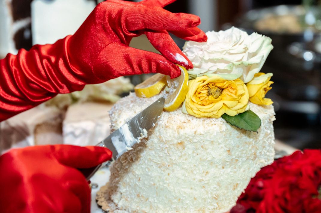 Hands wearing red gloves decorating a white cake with yellow and white roses.