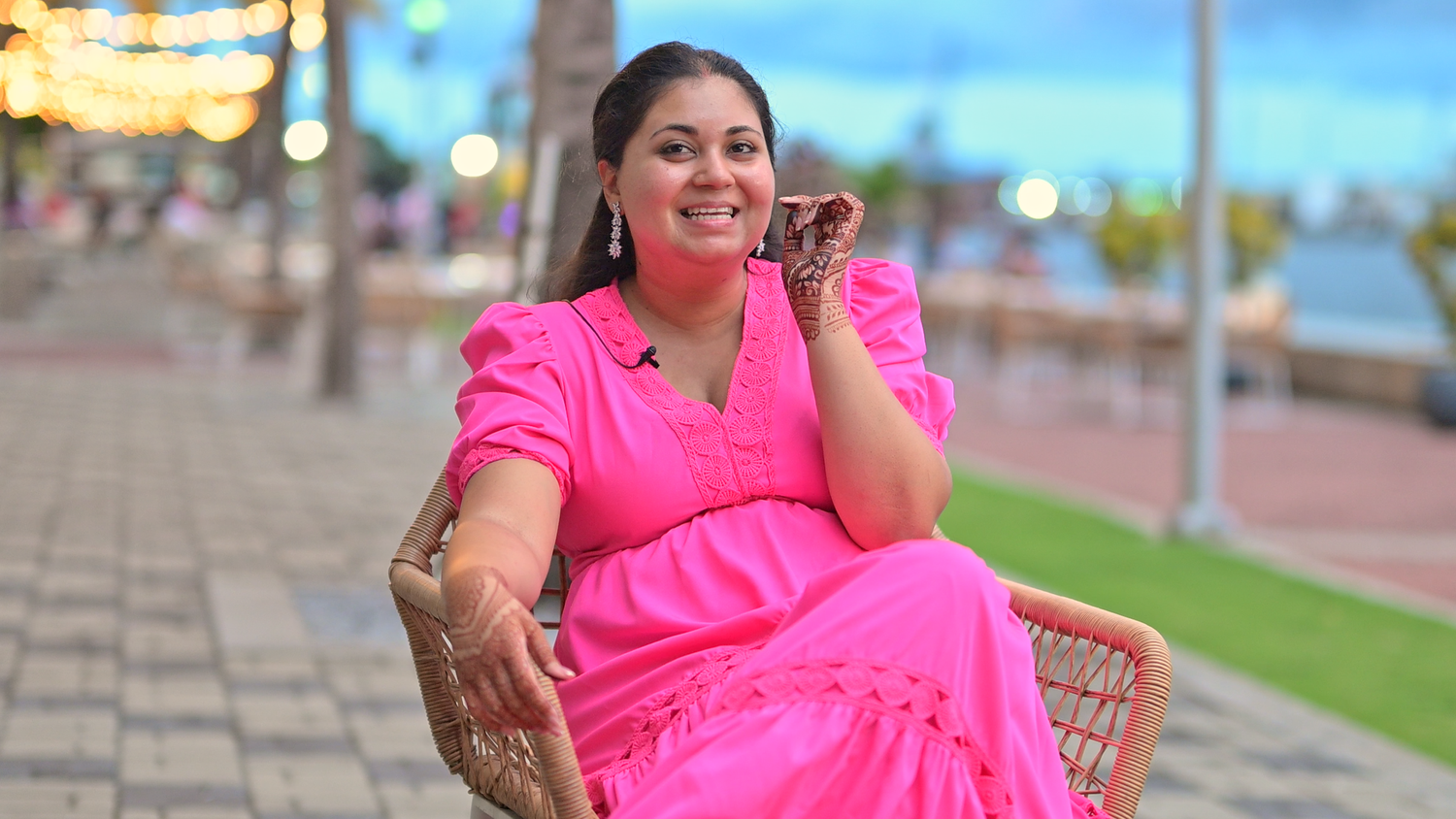 Woman in vibrant pink traditional dress smiling while seated outdoors at sunset beside a waterfront walkway.