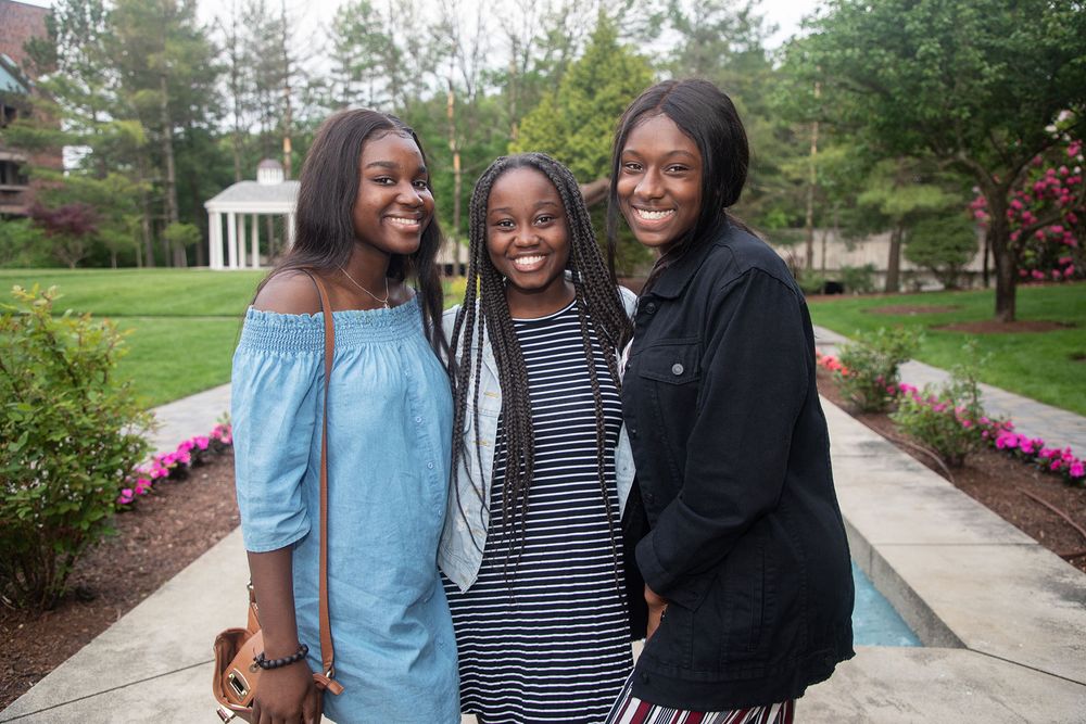 Three friends pose together on a stone pathway with flowering gardens and trees in the background.