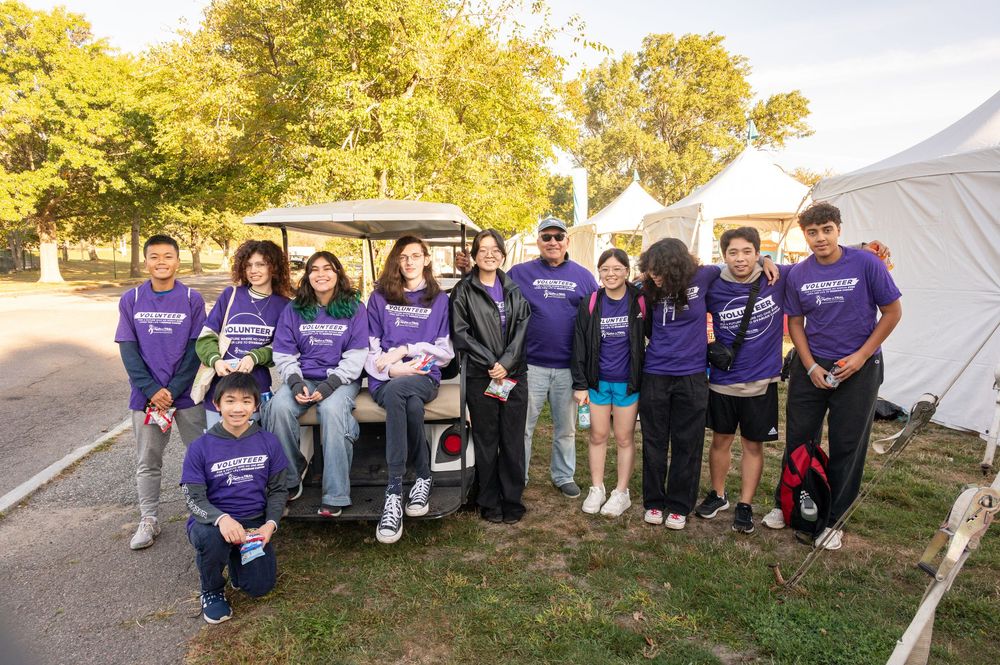 A group of volunteers in purple t-shirts gather outdoors near trees during a community event.