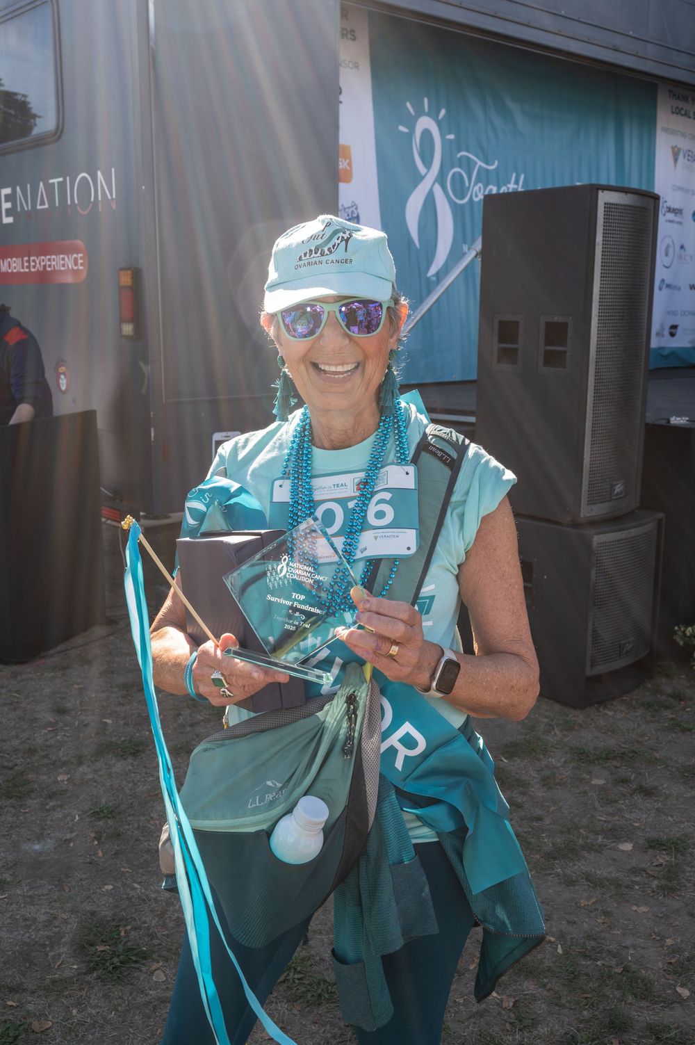 A volunteer wearing teal colored vest and sunglasses holds registration materials at an outdoor event.