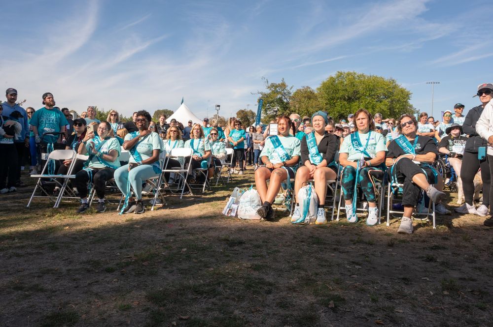 A large crowd of people in turquoise shirts gather outdoors on a sunny day for a group event.