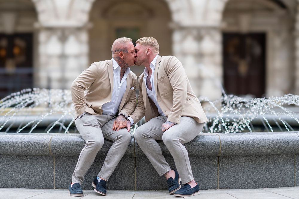 Two people in coordinating beige suits lean close together while sitting on a fountain ledge with ornate architecture behind them.