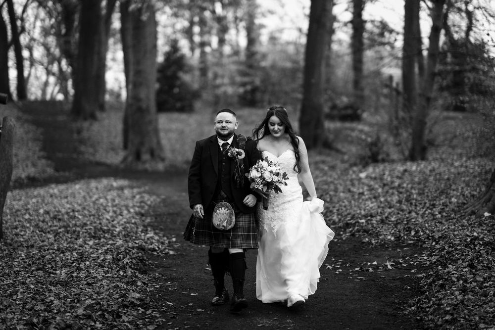 A couple walks down a wooded path at their traditional Scottish wedding, with the groom in a kilt and bride in white.